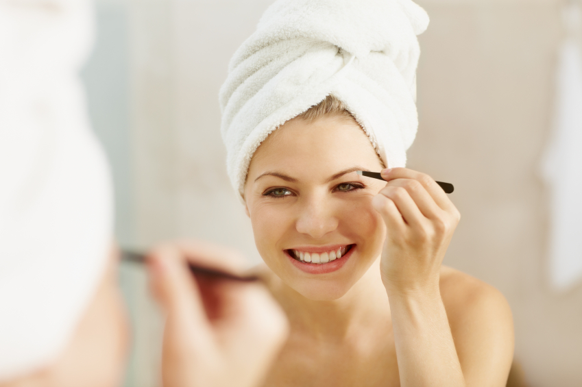 Beautiful young woman smiling while looking in her bathroom mirror plucking her eyebrows
